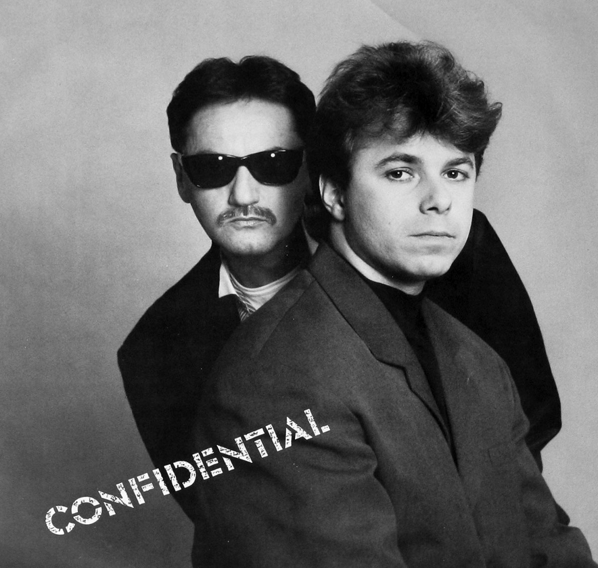 Custom inner sleeve photo of the 1989 Special EFX album Confidential, GRP Records. The image is a black and white studio portrait of George Jinda and Chieli Minucci. Jinda, on the left, wears dark sunglasses, a mustache, and a dark jacket with a serious, enigmatic look. Minucci, in front to the right, wears a tailored suit jacket and black turtleneck, looking directly into the camera with a calm yet intense expression. The background is plain grey, focusing all attention on the duo. The word Confidential appears in bold stencil-style letters slanted diagonally across the lower part of the image, reinforcing the album’s theme of secrecy and intrigue.
